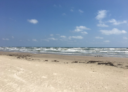 A sandy beach with gentle waves, under a clear blue sky with a few clouds.