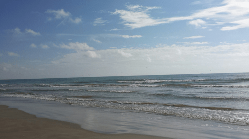 A serene beach scene with gentle waves, a clear blue sky, and soft clouds reflecting on the water.