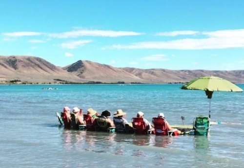 A group of people sitting in shallow water on beach chairs under an umbrella, with mountains in the background.
