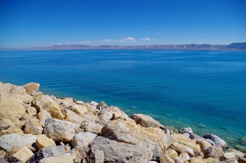 A serene view of a calm blue lake with rocky shores and distant mountains under a clear blue sky.