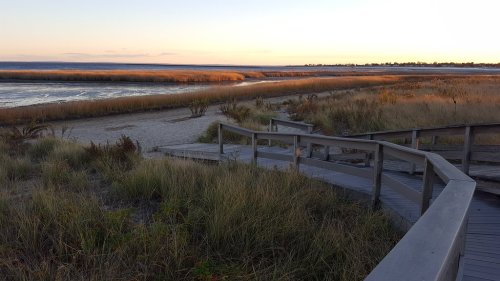 A wooden walkway leads through tall grass to a serene beach at sunset, with calm waters in the background.