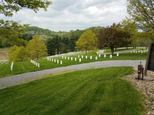 A serene cemetery with white headstones arranged in rows, surrounded by green grass and trees under a cloudy sky.