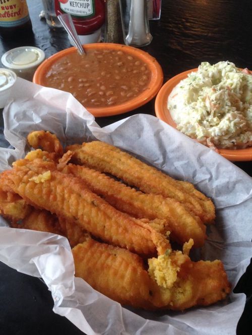 A basket of golden fried fish fillets with sides of coleslaw and baked beans on a table.