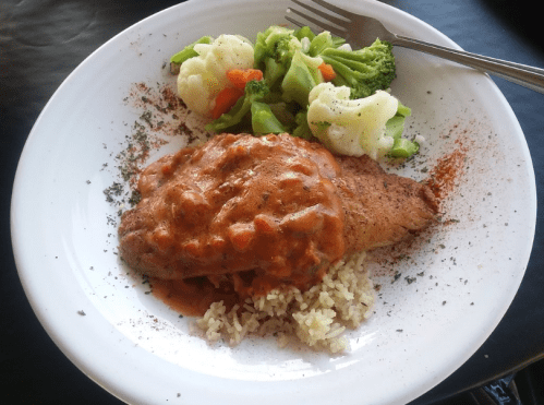 A plate of seasoned fish topped with a tomato sauce, served with rice and a side of mixed steamed vegetables.