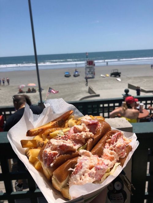 A plate of lobster rolls and fries with a beach view in the background on a sunny day.