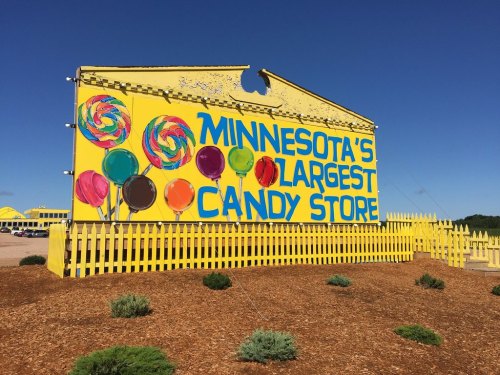 Bright yellow sign for "Minnesota's Largest Candy Store" with colorful lollipops and a blue sky in the background.