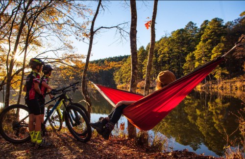 A person relaxes in a hammock by a lake, while two kids with bikes enjoy the scenic autumn landscape.