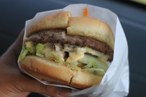 A hand holding a cheeseburger with lettuce and onion, wrapped in white paper.