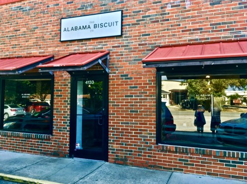 Exterior of The Alabama Biscuit Co. with a brick facade and red awning, featuring large windows and a welcoming entrance.