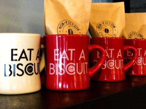 Four coffee mugs with the phrase "EAT A BISCUIT," alongside a bag of coffee on a shelf.