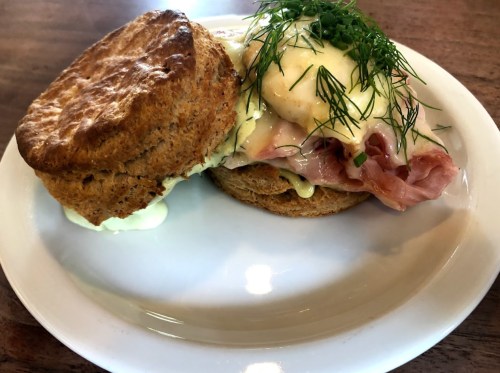 A biscuit sandwich with ham, poached egg, hollandaise sauce, and fresh herbs on a white plate.