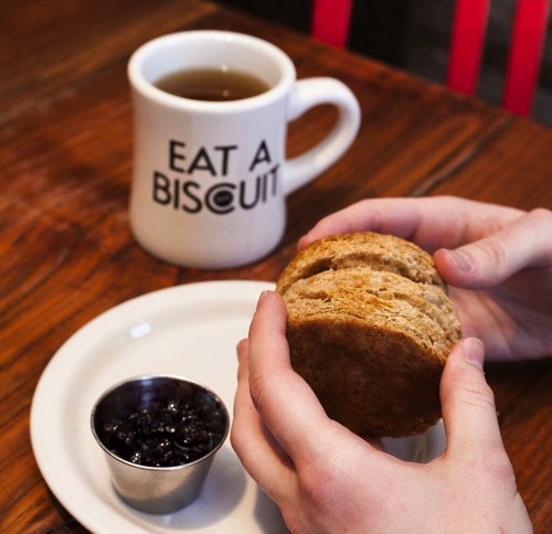 A person holds a biscuit next to a cup of tea and a small dish of jam on a wooden table.