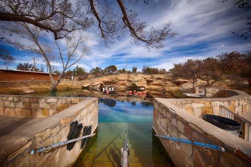 A serene natural pool surrounded by stone steps, trees, and a clear blue sky, with colorful buoys floating in the water.