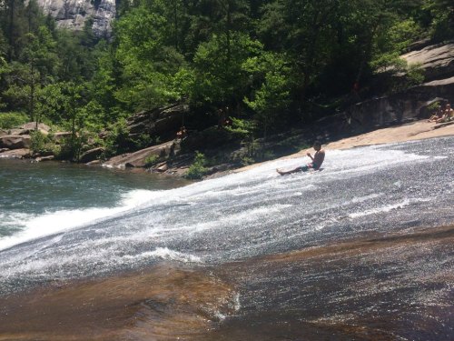 A person slides down a smooth rock into a river, surrounded by lush greenery and rocky terrain.