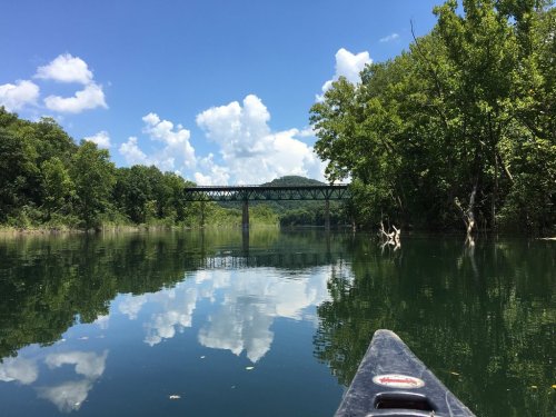A serene river scene with a bridge in the background, surrounded by lush greenery and reflecting clouds on the water.