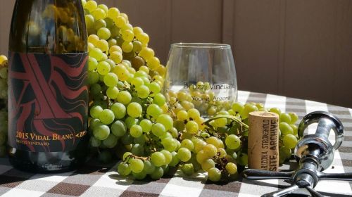 A bottle of 2015 Vidal Blanc wine, a glass, grapes, a cork, and a corkscrew on a checkered tablecloth.