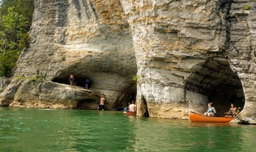 Two canoes near rocky cliffs with caves, surrounded by clear green water and lush greenery.