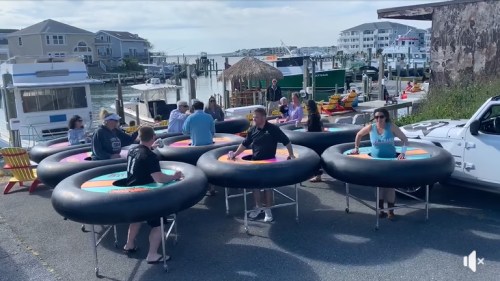 A group of people enjoying a sunny day on inflatable trampolines by a marina, with boats and buildings in the background.