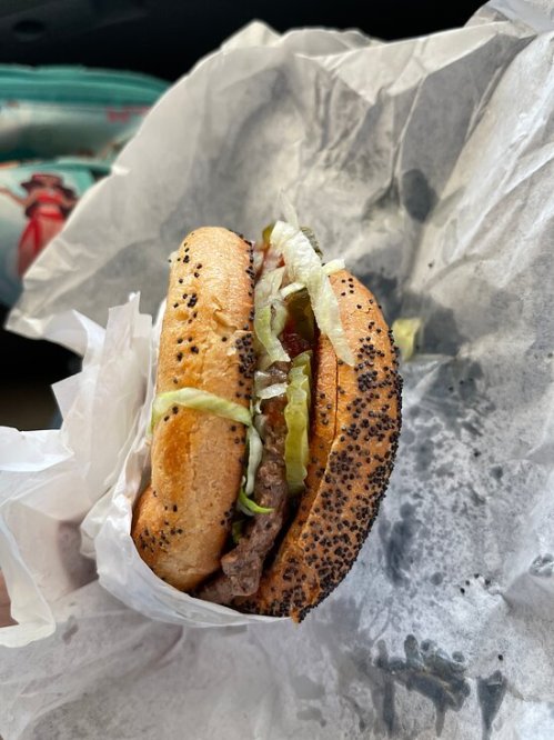 A close-up of a poppy seed bun sandwich filled with lettuce, meat, and condiments, wrapped in white paper.