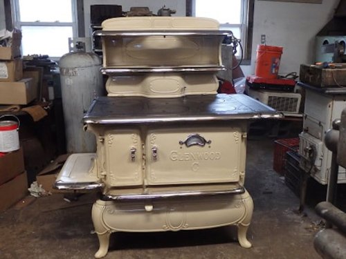 A vintage cream-colored stove with metal accents, featuring an ornate design and multiple compartments, in a cluttered workshop.