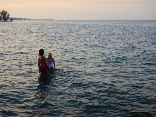 Two people wade in calm water during sunset, with a serene landscape in the background.