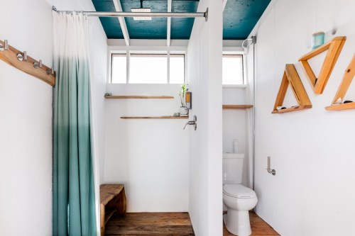 Bright, modern bathroom with a wooden bench, white walls, teal ceiling, and open shelving.