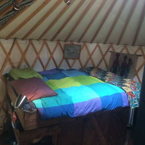Cozy yurt interior featuring a bed with colorful blankets and pillows, surrounded by wooden walls and soft lighting.