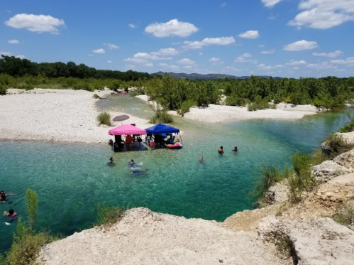 People enjoying a sunny day by a clear river, with colorful umbrellas and lush greenery in the background.