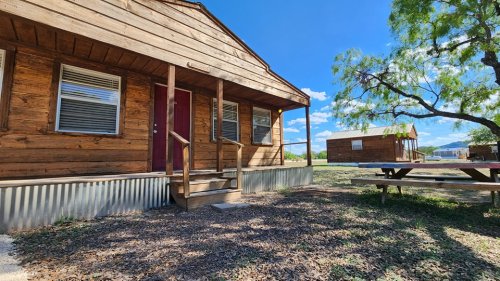 A wooden cabin with a red door, steps, and picnic table, surrounded by grass and trees under a blue sky.