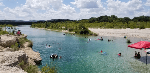 A scenic river with people swimming and floating, surrounded by greenery and a bright blue sky with fluffy clouds.