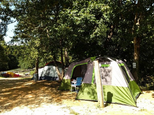 Two camping tents under trees by a river, with a blue chair and kayaks visible in the background.
