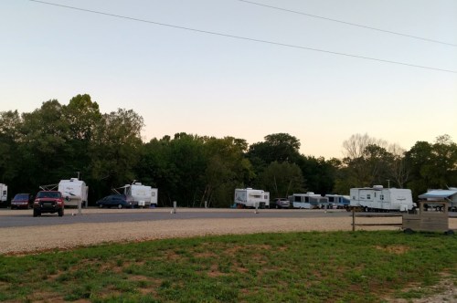 A campsite with several RVs parked along a gravel area, surrounded by trees and a clear sky at dusk.
