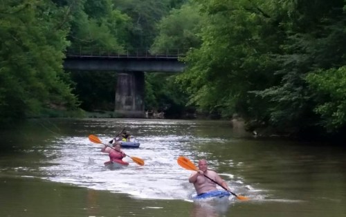 Two people kayak on a calm river surrounded by lush greenery, with a bridge visible in the background.