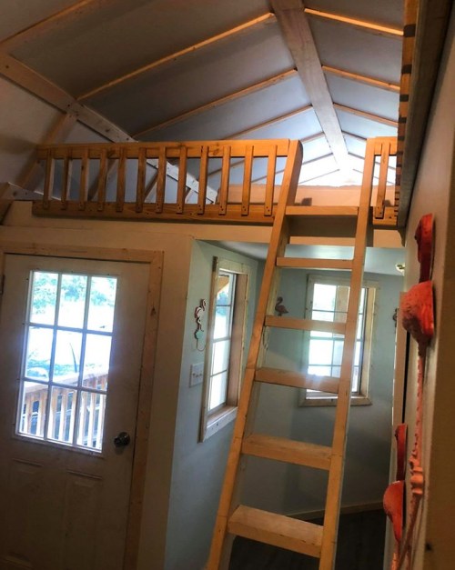 Interior of a small cabin with a loft, wooden ladder, and large windows, featuring a cozy, rustic design.