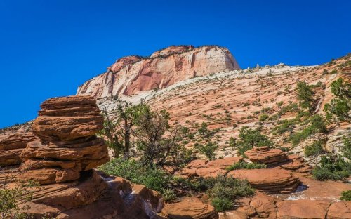 A rocky landscape with layered red and white cliffs under a clear blue sky, featuring scattered vegetation.