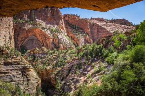 A scenic view of rugged red rock formations and lush greenery in a canyon, framed by a rocky overhang.