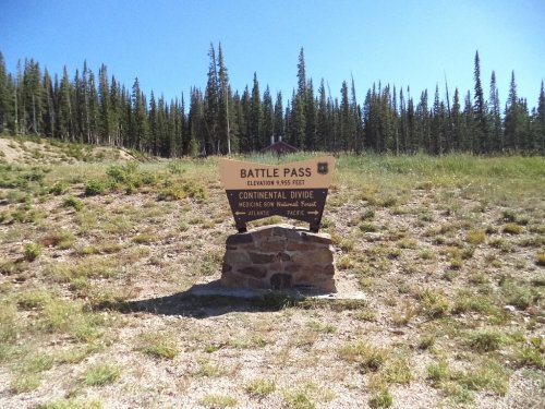 Sign at Battle Pass, marking the Continental Divide, surrounded by grassy terrain and evergreen trees. Elevation: 9,155 feet.