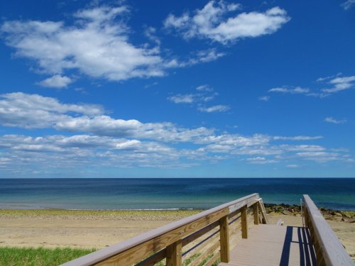 A wooden boardwalk leads to a serene beach with calm blue waters and a bright sky filled with fluffy clouds.