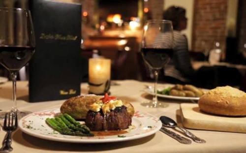 A gourmet meal featuring filet mignon, asparagus, a baked potato, and a bread roll, with wine glasses in a cozy restaurant setting.