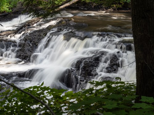 A serene waterfall cascades over dark rocks, surrounded by lush green foliage and trees.