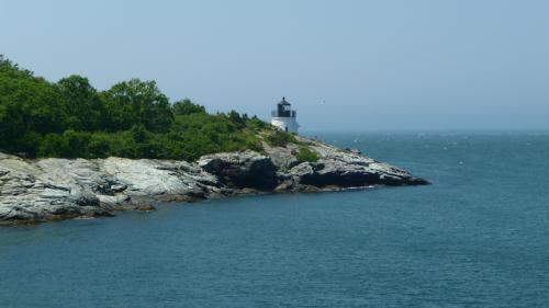 A lighthouse stands on rocky cliffs by the ocean, surrounded by greenery under a clear blue sky.