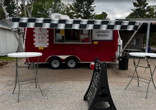 A red food truck with a black and white checkered awning, serving Lake Superior whitefish, with outdoor tables nearby.