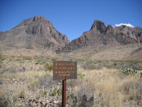 A trail sign points towards various hiking paths in a rugged desert landscape with mountains in the background.