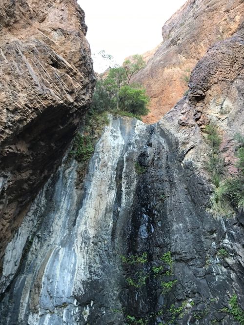 A rocky cliff with vertical layers, featuring a small waterfall and sparse greenery at the top.