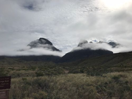 Clouds hover over rugged mountains, surrounded by green vegetation and a signpost in the foreground.