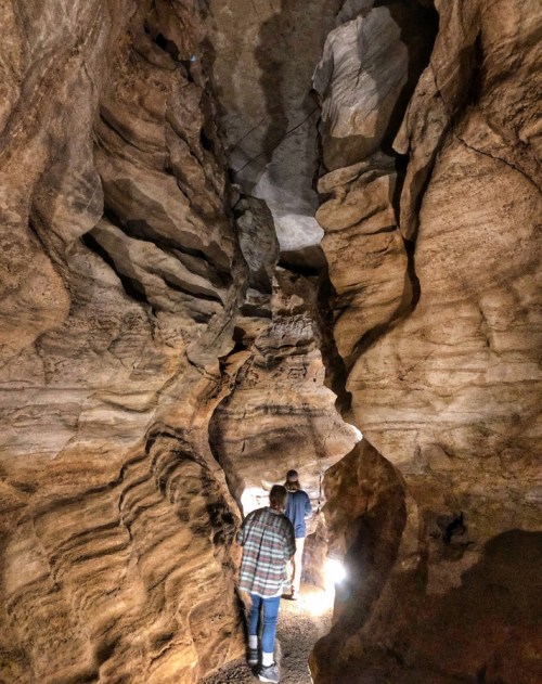 Two people walk through a narrow, winding cave with textured rock walls and soft lighting illuminating the path.