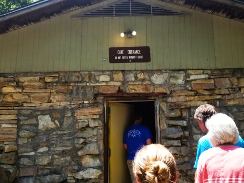 A group of people approaches a stone cave entrance with a sign that reads "Cave Entrance - Do Not Enter Without Guide."