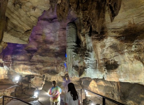 Two people explore a dimly lit cave with unique rock formations and colorful lighting.