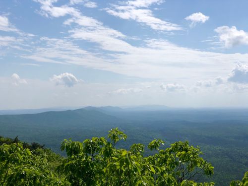 A panoramic view of rolling green mountains under a partly cloudy sky. Lush foliage in the foreground.