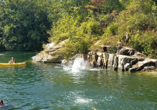 A sunny scene at a lake with people swimming, kayaking, and jumping off rocks into the water surrounded by trees.